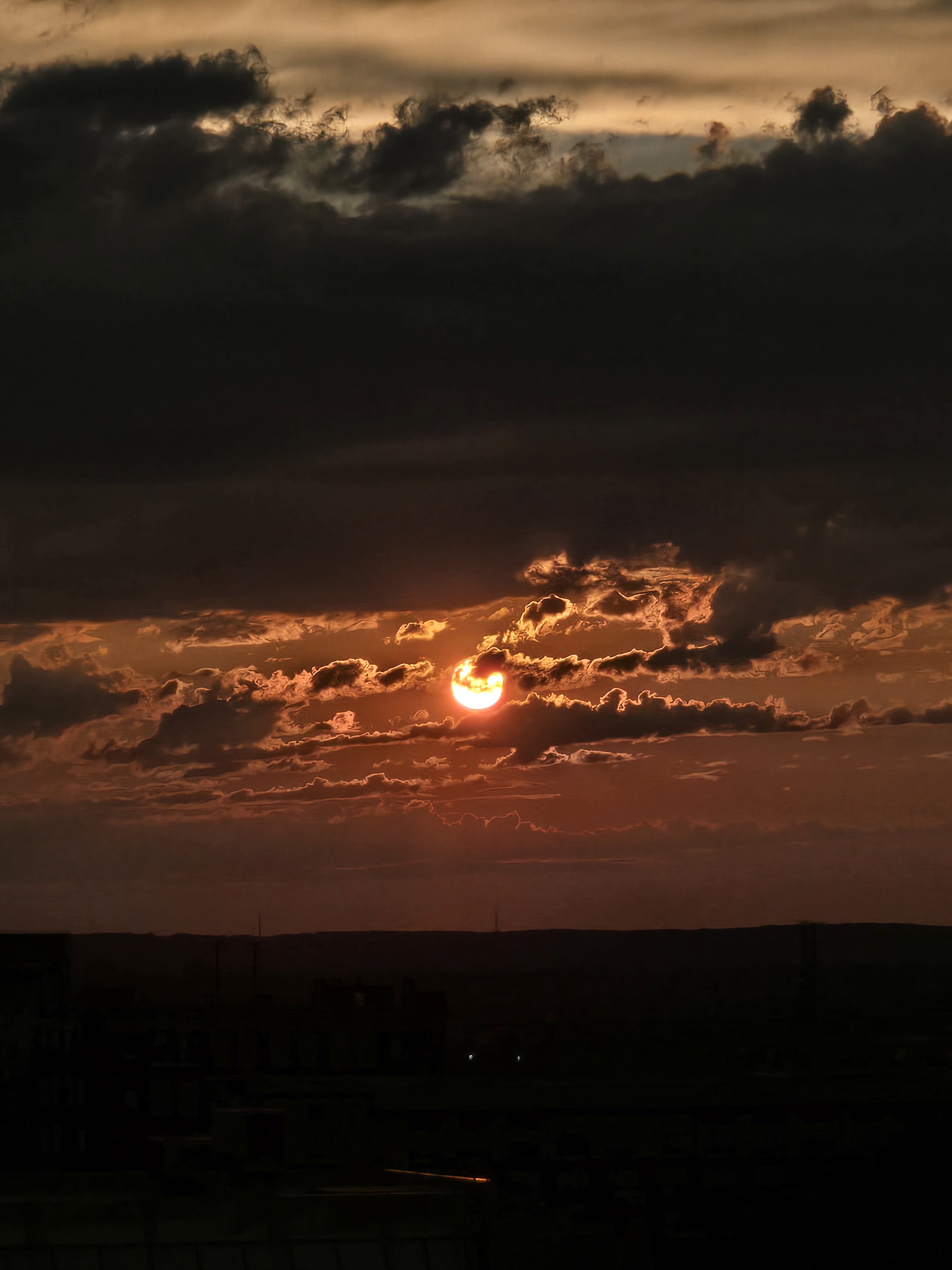 Dramatic sunset with the sun peeking through dark clouds above a city skyline